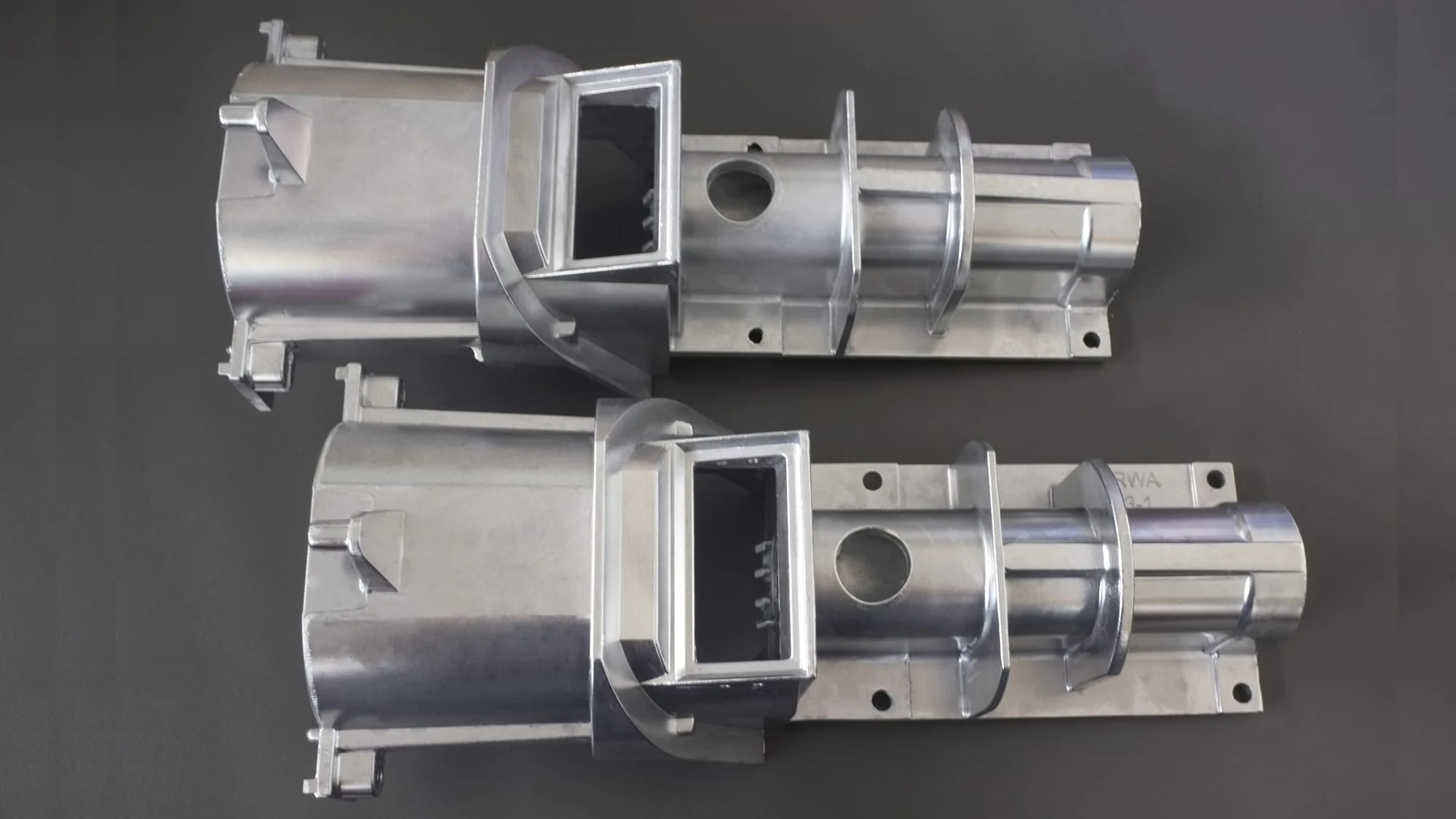 The image shows a pair of aluminum die casting parts on a gray background. The parts are made of metal and have a metallic sheen. They are arranged in a symmetrical pattern, with the largest part in t