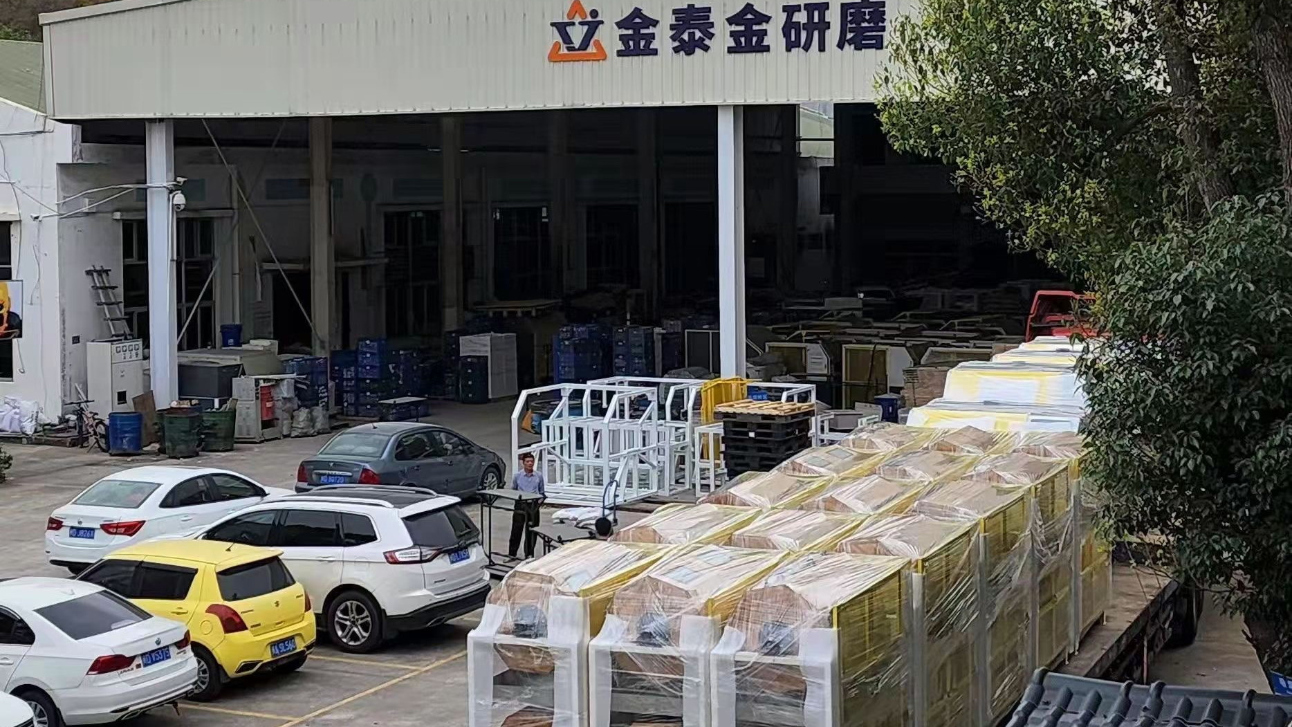 The 金泰金研磨 sign over the gate of the plant at No. 31 Xinchang Road, Xiamen, with machinery bays behind it and crated machines on the apron